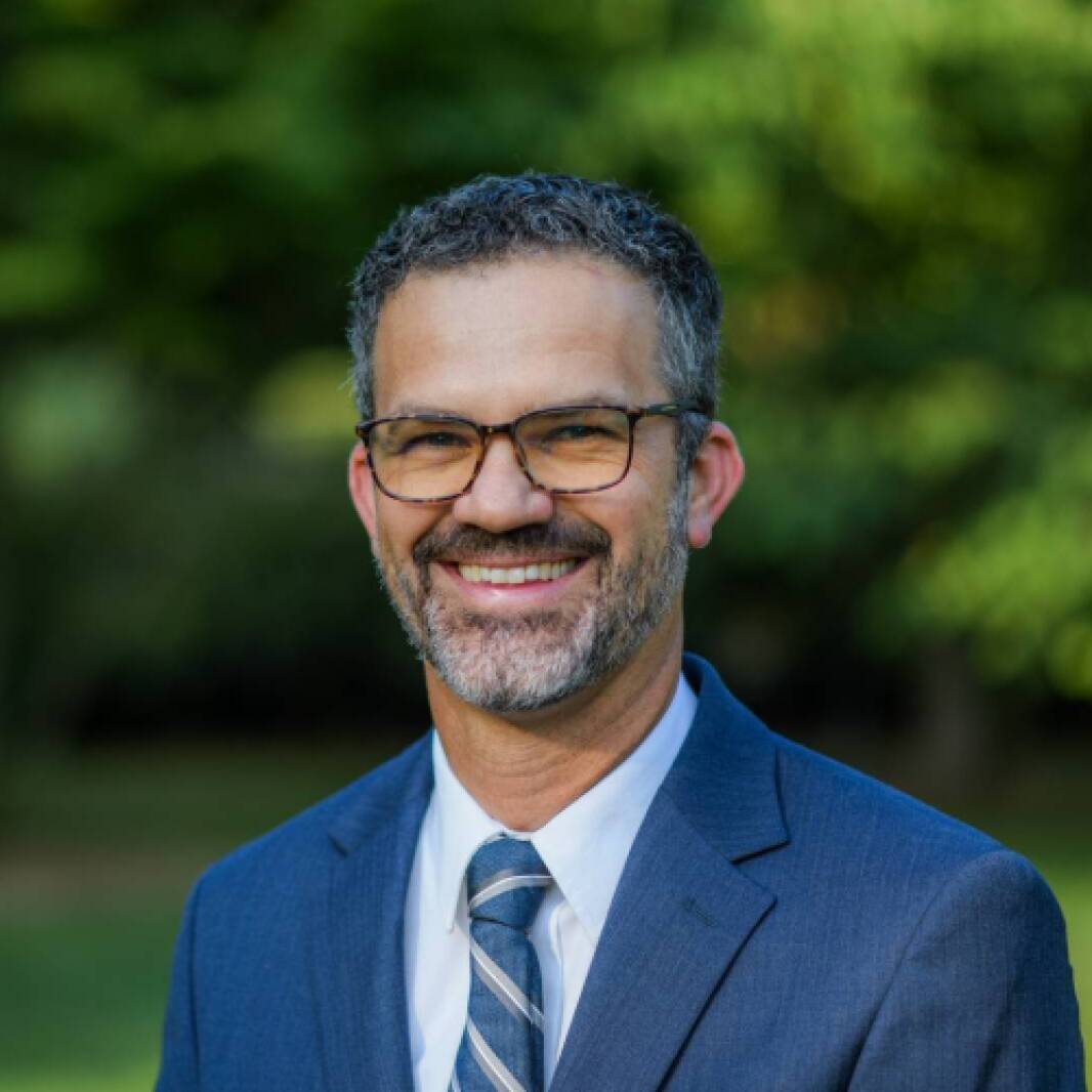 A man with dark hair, a gray beard, and black glasses wearing a blue blazer, shirt, and tie smiling in front of a green grass background